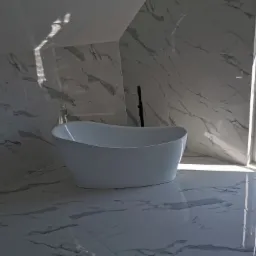 Wolnostojąca biała wanna w nowoczesnej łazience na poddaszu, wykończonej płytkami imitującymi marmur, z oknem wychodzącym na zieleń.