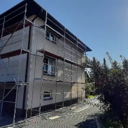 Dwupiętrowy dom w trakcie prac elewacyjnych, osłonięty rusztowaniem, z widocznym fragmentem zaparkowanego samochodu dostawczego po lewej stronie i intensywnym słońcem w tle.