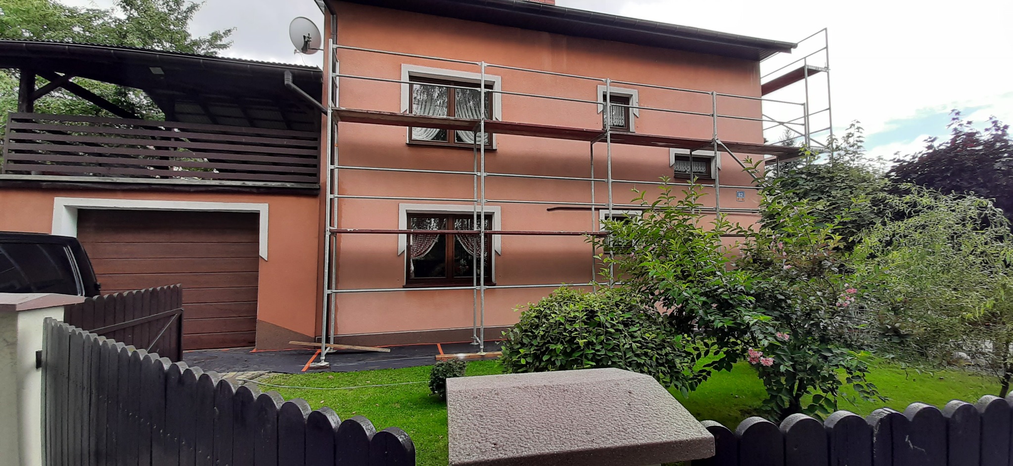 Dom z różową elewacją w trakcie malowania, widoczne rusztowania przy oknach z firankami, tło z zielenią i drewnianym garażem.