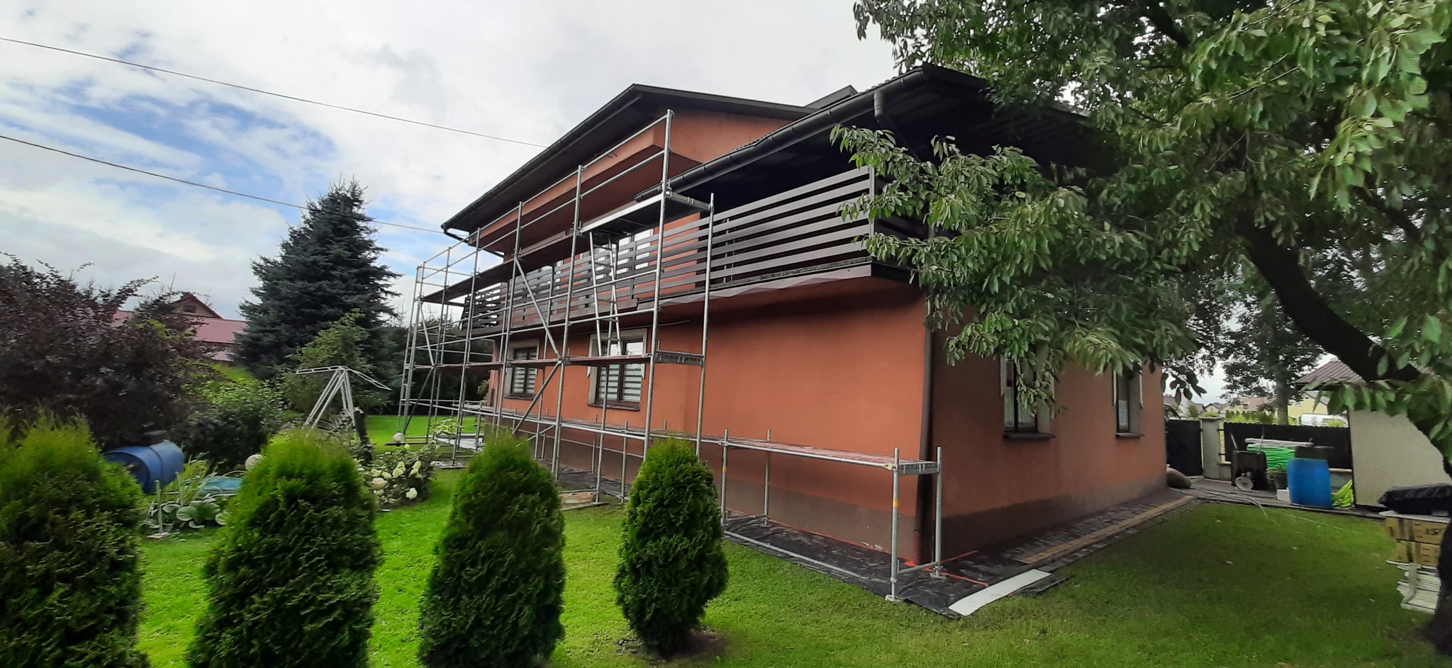 Dom jednorodzinny z elewacją w odcieniach terakoty, otoczony zielenią i częściowo zasłonięty rusztowaniem podczas prac elewacyjnych.