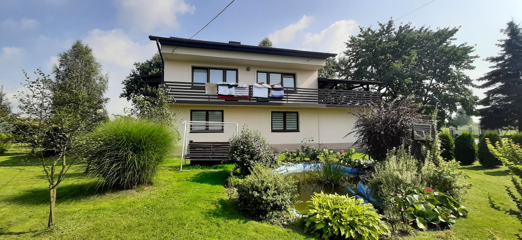 Dwupiętrowy dom z beżową elewacją i ciemnymi oknami, balkonem z suszącym się praniem, otoczony zielonym ogrodem z oczkiem wodnym i różnorodną roślinnością.