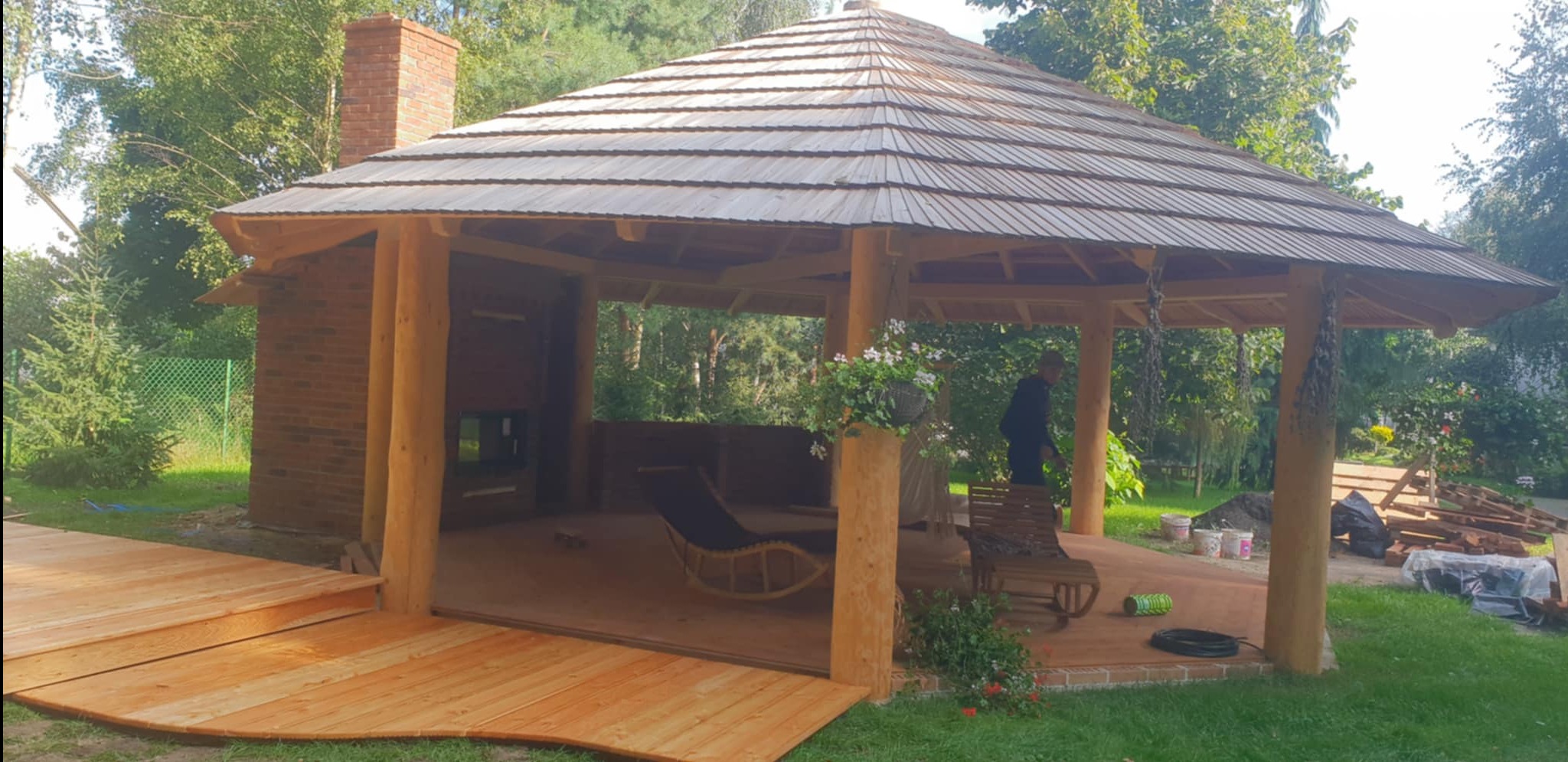 Zadaszona konstrukcja z drewnianymi słupami i dachem z drewnianych desek, przylegająca do ceglanej ściany z kominkiem, z drewnianym tarasem i meblami ogrodowymi wewnątrz.