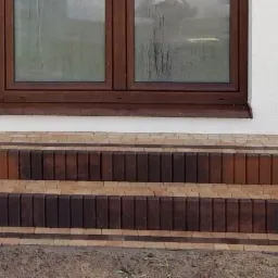 Schody wejściowe do domu wyłożone kostką brukową w odcieniach brązu i beżu, z widocznymi detalami wzoru i fug, na tle białej ściany budynku z brązowymi oknami.