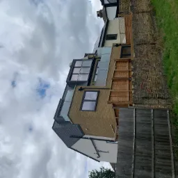 Murowany dom z nowoczesnymi oknami i szklanymi balkonami, widok z dołu na tle pochmurnego nieba, obok mur z cegły i drewniany płot.