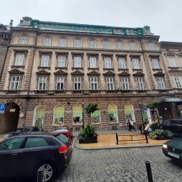 Gabinety Psychoterapii ARIADNA Bielsko-Biała - Elewacja kamienicy z oknami i szyldami reklamowymi w oknach na parterze, zaparkowane samochody, brukowa ulica.