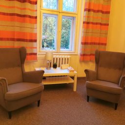 Gabinety Psychoterapii ARIADNA Bielsko-Biała - Gabinet terapeutyczny z dwoma fotelami typu uszak, stolikiem kawowym, lampką nocną i oknem z widokiem na zieleń.