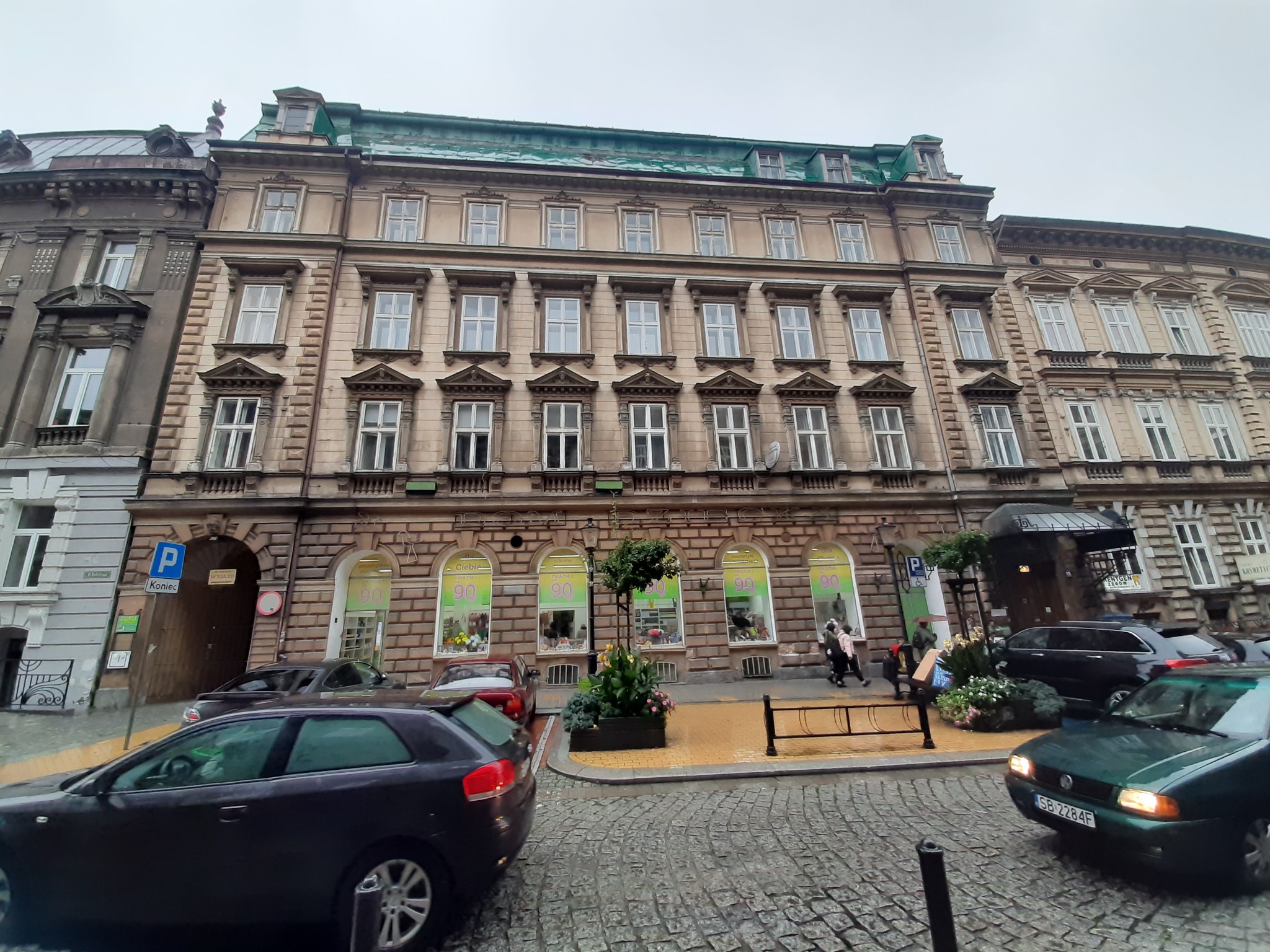 Elewacja kamienicy z oknami i szyldami reklamowymi w oknach na parterze, zaparkowane samochody, brukowa ulica.