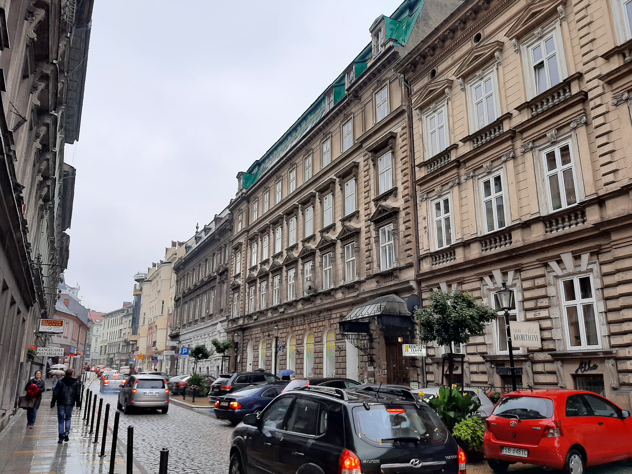 Ulica w europejskim mieście z kamienicami, samochodami i pieszymi w pochmurny dzień. Widoczny szyld 'Alba Kosmetyczny'