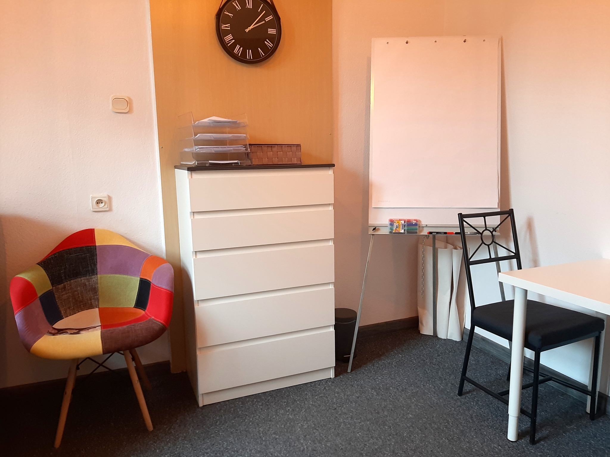 Jasne wnętrze gabinetu z komodą, zegarem ściennym, flipchartem, biurkiem i krzesłem patchworkowym, tworzące przyjazną atmosferę.