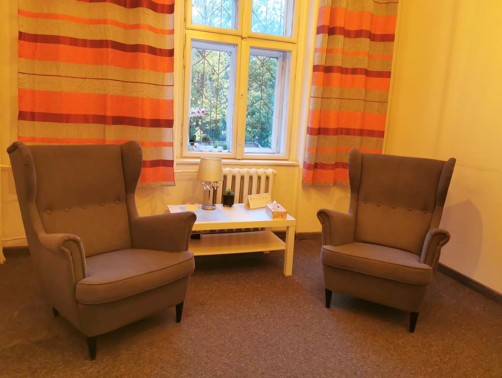 Gabinet terapeutyczny z dwoma fotelami typu uszak, stolikiem kawowym, lampką nocną i oknem z widokiem na zieleń.