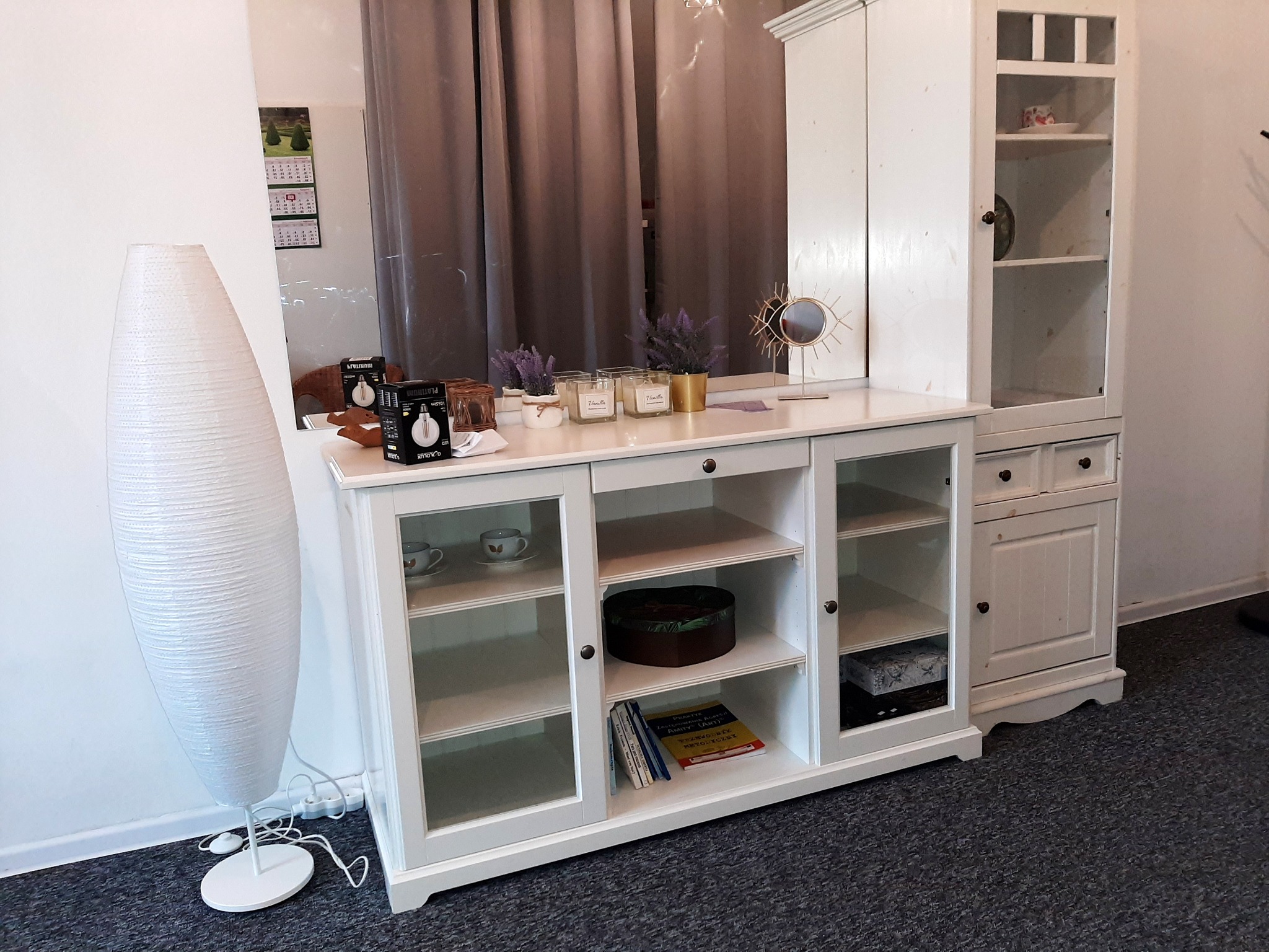 Jasny pokój z białymi meblami: kredens z szklanymi drzwiczkami, szafka z półkami i wysoka lampa podłogowa z papieru ryżowego. Na kredensie dekoracje, w tym świece i lusterko.