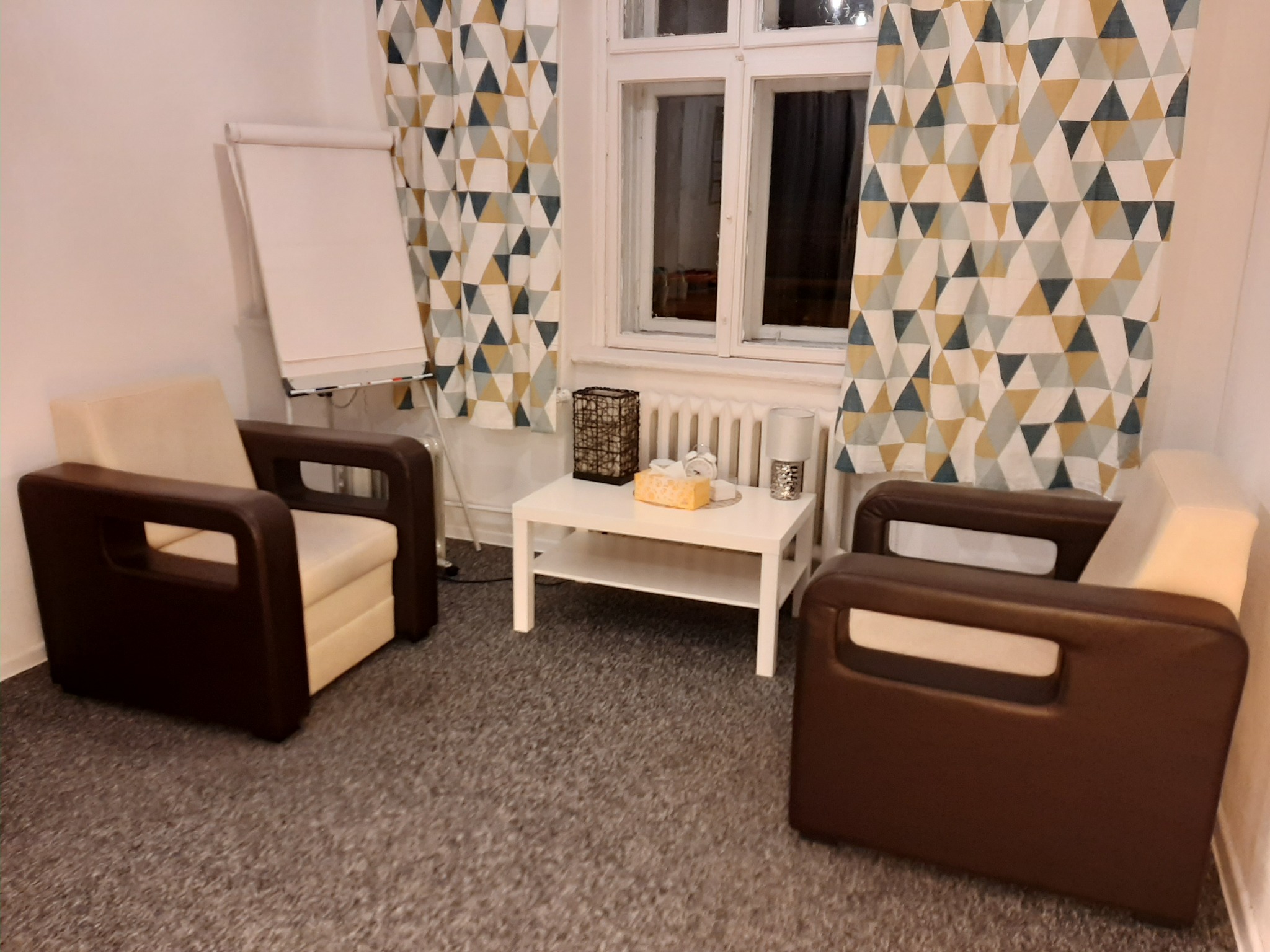 Pokój z dwoma fotelami z brązowymi bokami i kremowymi siedziskami, biały stolik kawowy z lampką i pudełkiem chusteczek, flipchart przy ścianie i zasłony w geometryczne wzory.