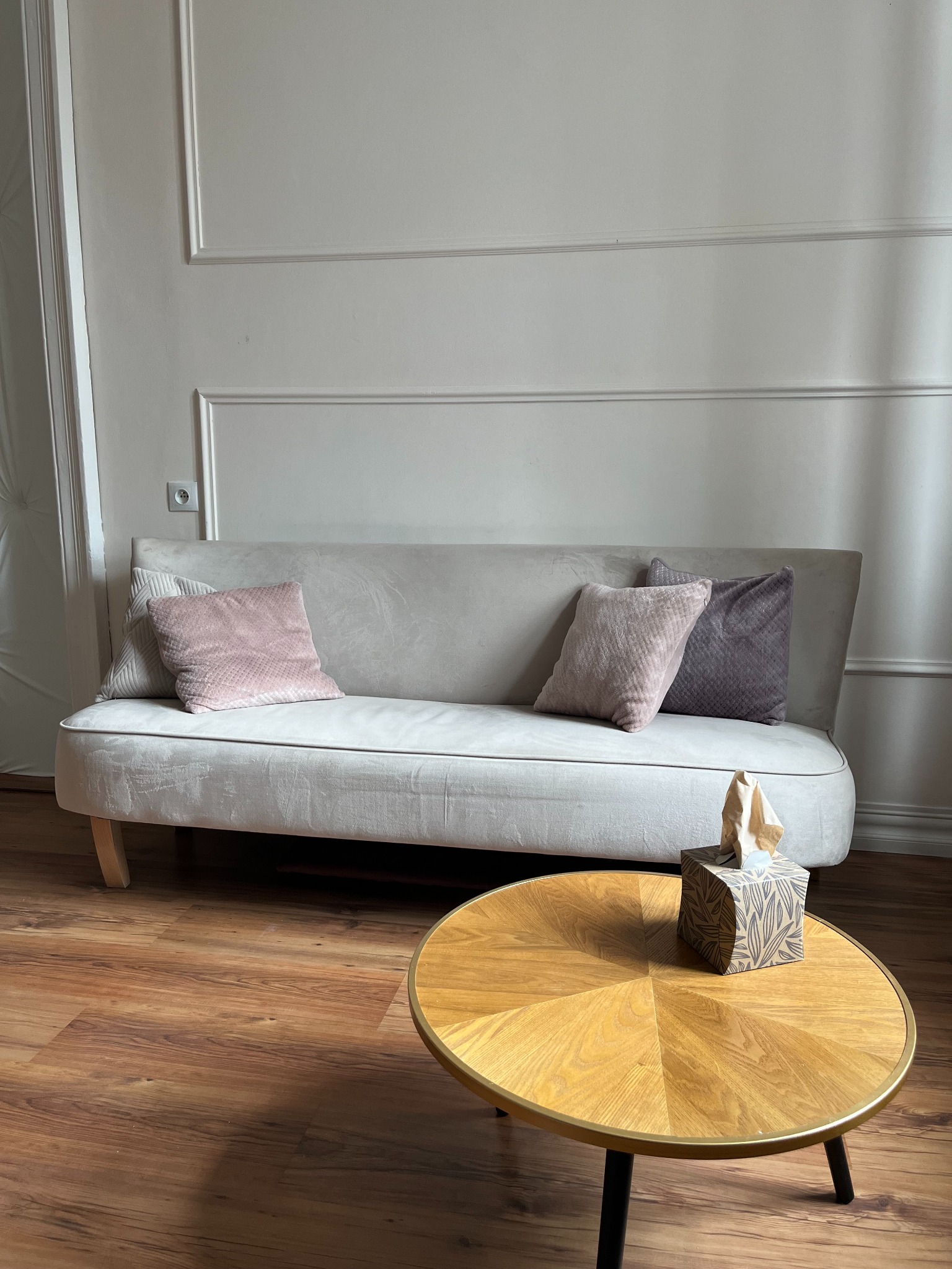 Jasnoszary tapczan z trzema poduszkami w odcieniach różu i fioletu, obok okrągły stolik z pudełkiem chusteczek, na podłodze panele imitujące drewno.