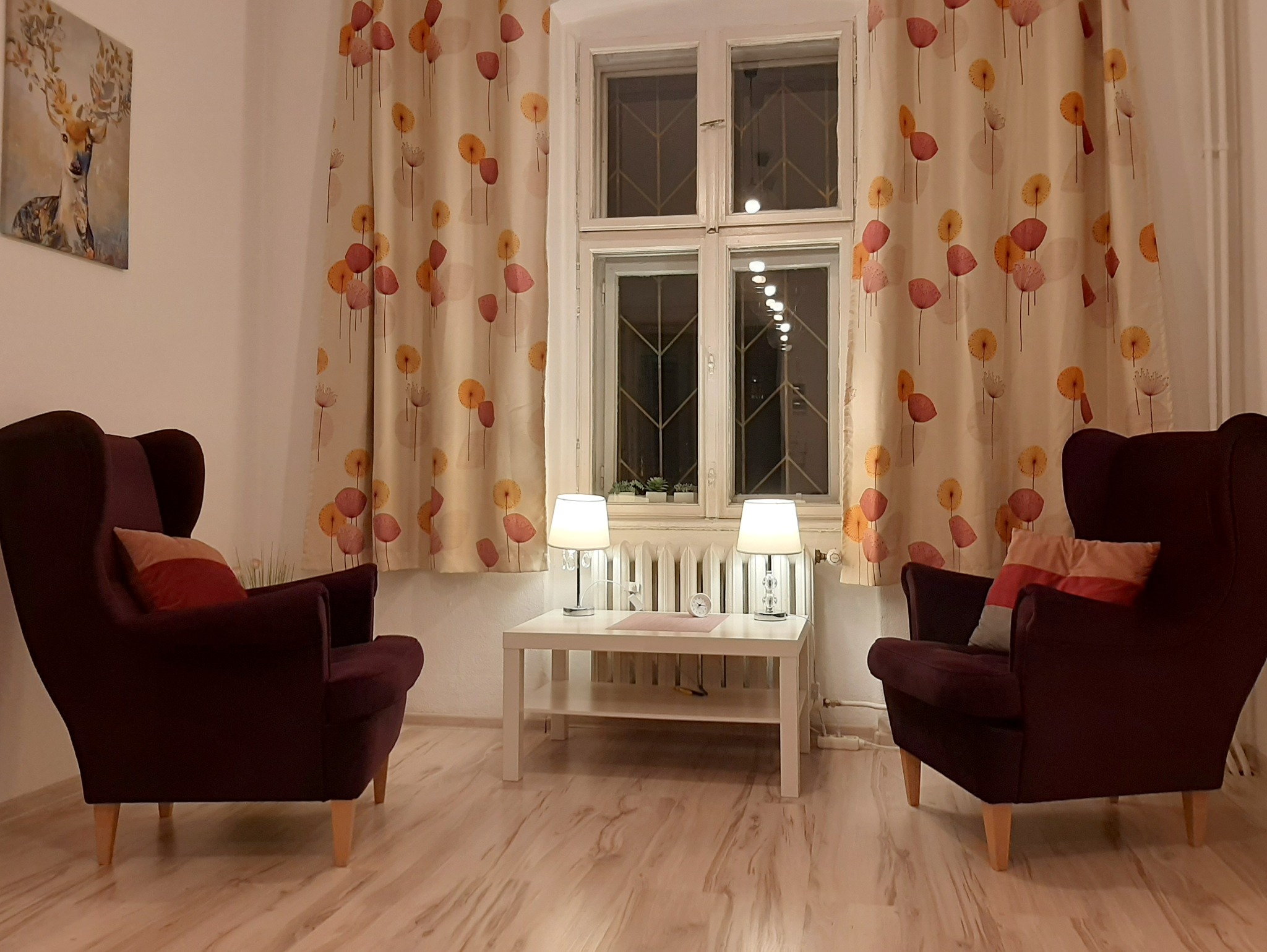 Gabinet terapeutyczny z dwoma bordowymi fotelami typu uszak, białym stolikiem z lampkami i obrazem jelenia na ścianie, tworzący przytulną atmosferę.