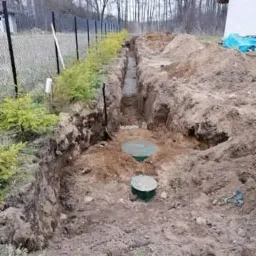 Wykopany rów z betonowym dnem i dwa zielone włazy szamba betonowego w ziemi, widok z góry na tle ogrodzenia z siatki i drzew.