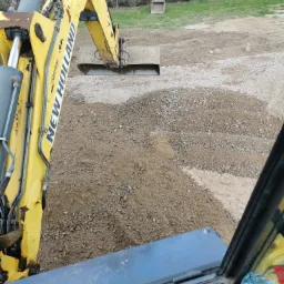 Żółta koparka New Holland, widziana z kabiny, podczas niwelacji terenu w Szczytnie, z pryzmą ziemi i gruzu przed łyżką.