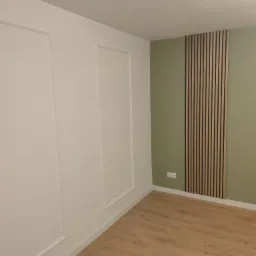 Wnętrze pokoju po remoncie: białe ściany z dekoracyjnymi ramkami, oliwkowa ściana z drewnianymi panelami pionowymi i jasna drewniana podłoga.