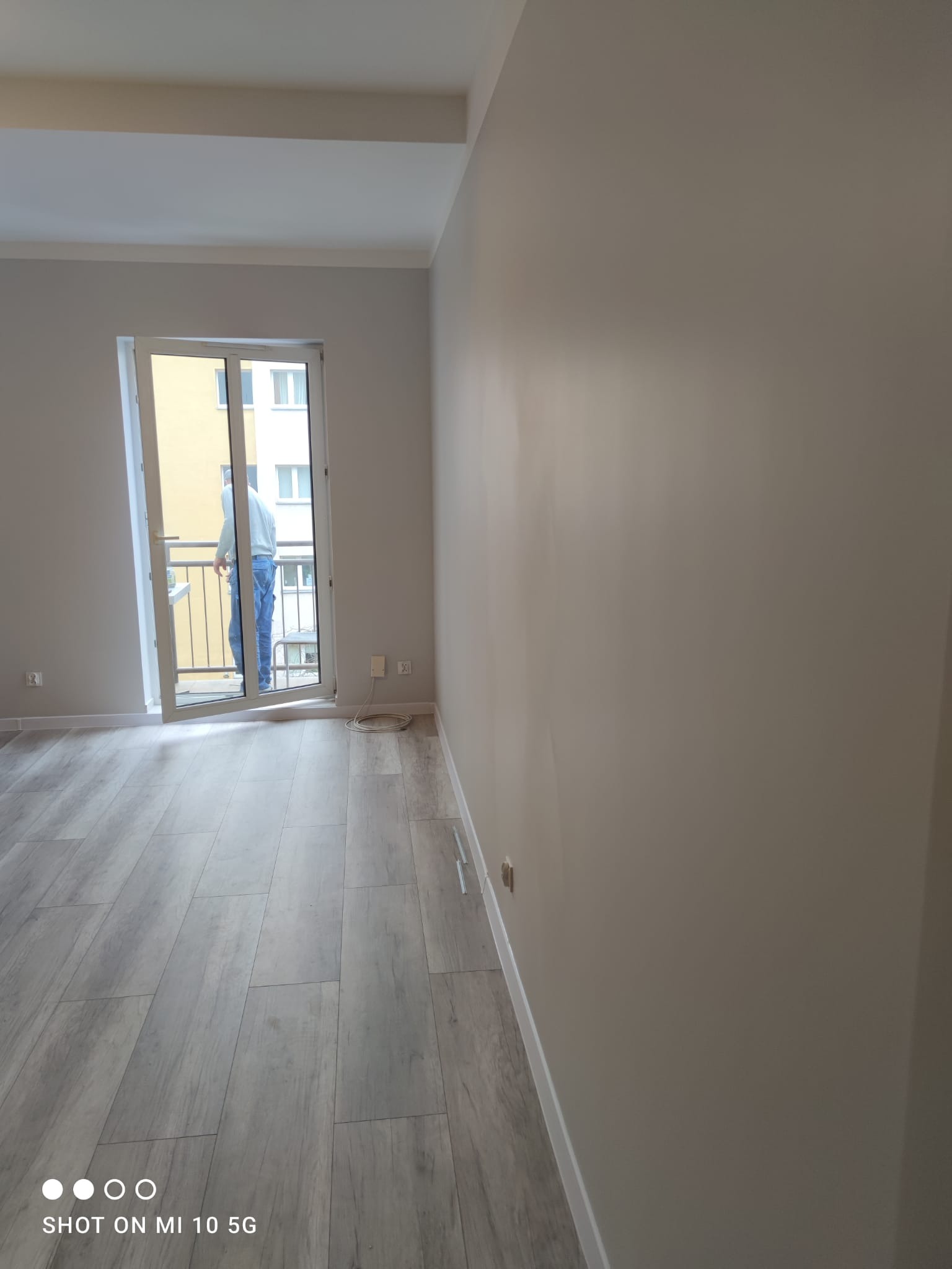 Wnętrze pokoju po remoncie z pomalowanymi ścianami na jasny odcień szarości i wykończonymi podłogami z paneli imitujących drewno; widoczne listwy przypodłogowe i otwarte drzwi balkonowe z widokiem...