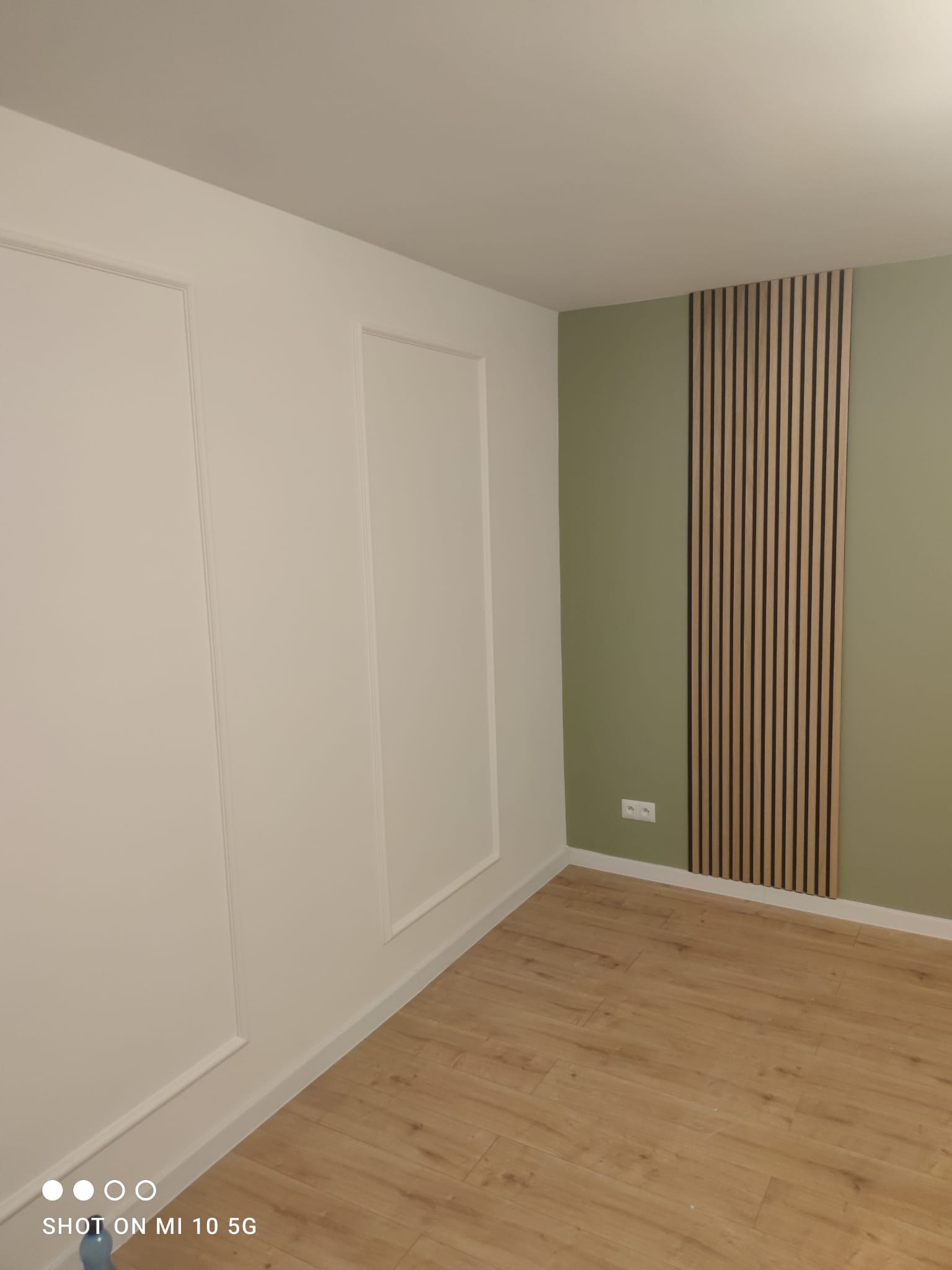 Wnętrze pokoju po remoncie: białe ściany z dekoracyjnymi ramkami, oliwkowa ściana z drewnianymi panelami pionowymi i jasna drewniana podłoga.