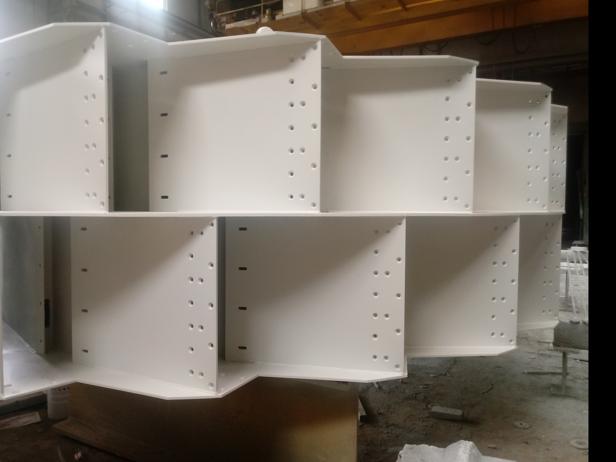 Białe, pomalowane proszkowo, stalowe elementy konstrukcyjne z otworami, ułożone w stos na podłodze w hali produkcyjnej.