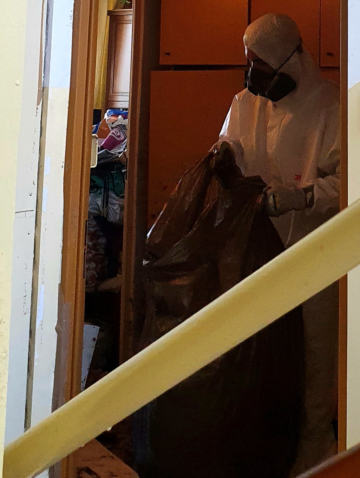 Osoba w kombinezonie ochronnym i masce trzyma duży, brązowy worek na śmieci w korytarzu mieszkania, widoczne schody i bałagan w tle.