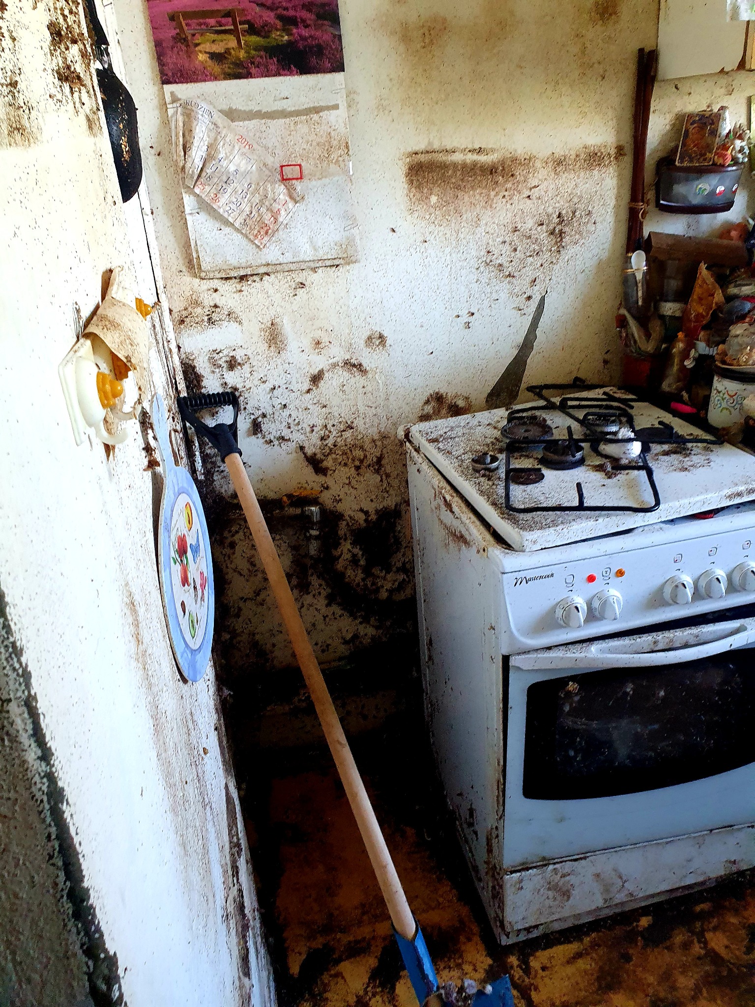 Zaniedbana kuchnia z brudnymi ścianami i kuchenką, łopata oparta o ścianę, widoczny stary kalendarz na ścianie.