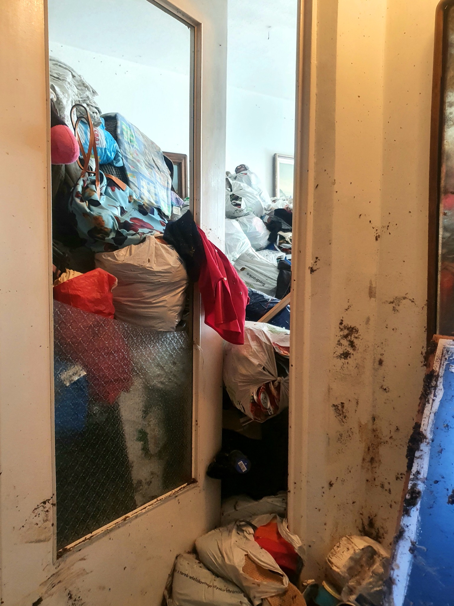 Zaniedbane pomieszczenie z widocznymi śladami zabrudzeń na ścianach i podłodze, wypełnione workami i różnymi przedmiotami, widoczne przez częściowo otwarte drzwi z matową szybą.