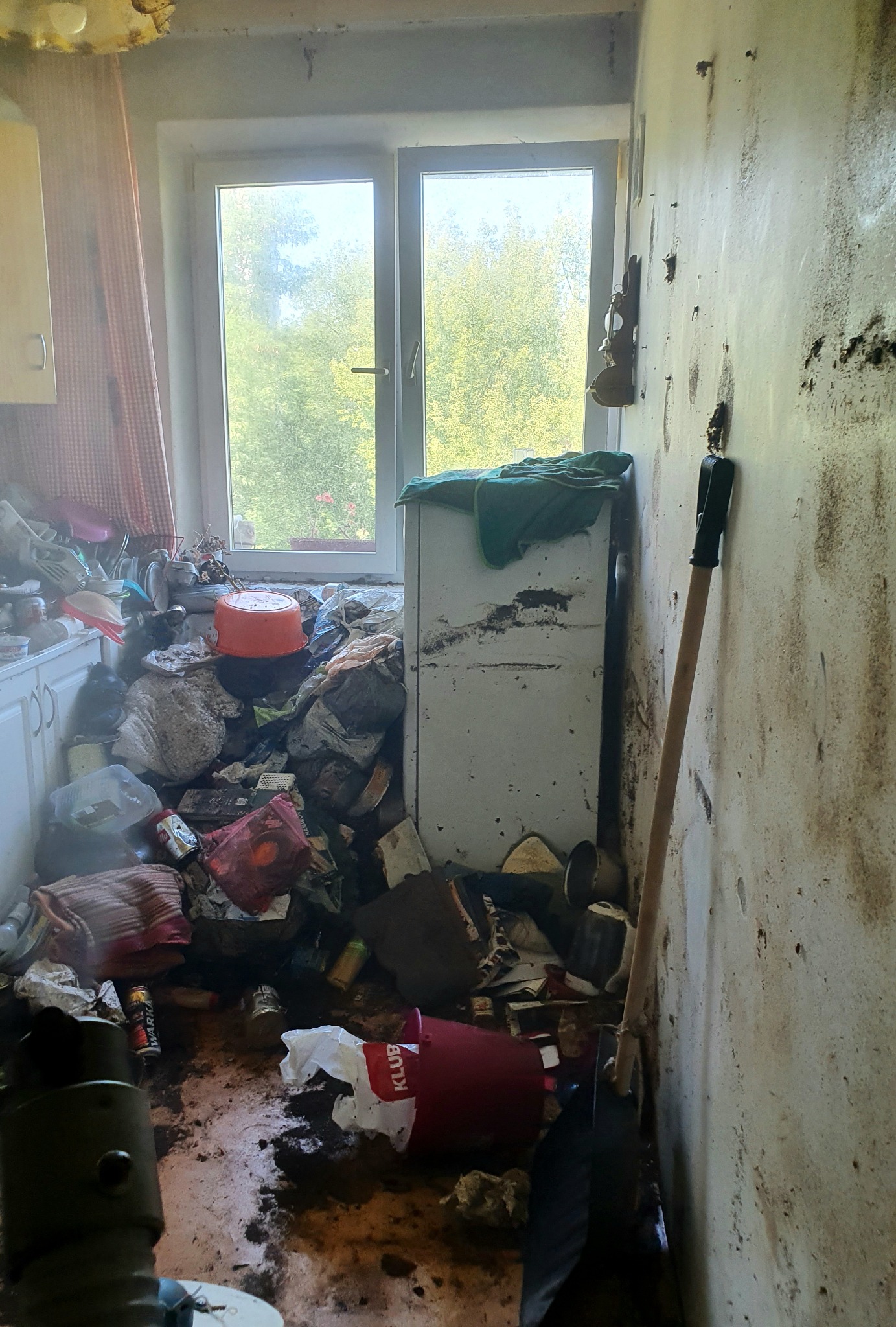 Zaniedbane wnętrze kuchni z widocznymi zabrudzeniami i nagromadzeniem śmieci, przy ścianie oparta łopata, okno z widokiem na zieleń na zewnątrz.