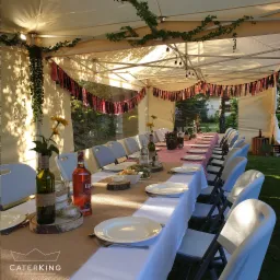 Uroczysty stół w stylu BOHO pod namiotem, dekorowany kwiatami, butelkami owiniętymi jutą i girlandami w kratę, nakryty białą zastawą i bieżnikiem z juty.