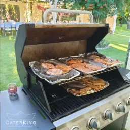 Grill gazowy z otwartą pokrywą, na ruszcie aluminiowe tacki z pieczonymi mięsami i kiełbasami, w tle trawnik, namiot cateringowy i przygotowane stoły, na boku grilla zapalona świeczka w szklanym...