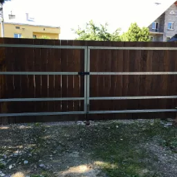 Dwuskrzydłowa brama wjazdowa z ciemnego drewna, wzmocniona metalowymi profilami, z widocznym zamkiem i zawiasami, osadzona pomiędzy ceglanymi słupkami na tle zabudowy mieszkalnej.