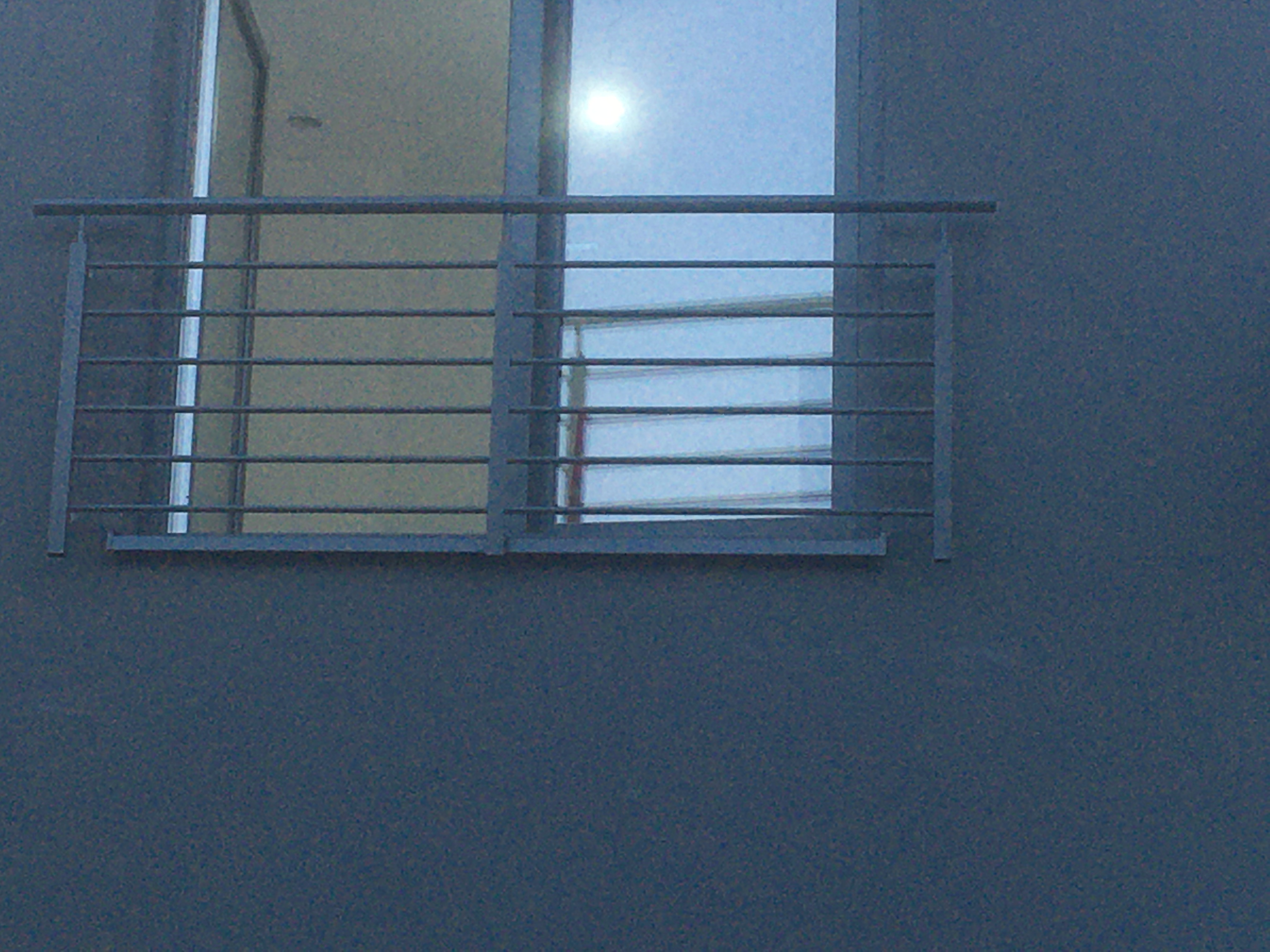 Minimalistyczna, metalowa barierka balkonowa z poziomymi szczeblami, zamontowana na szarym budynku, widok z zewnątrz.