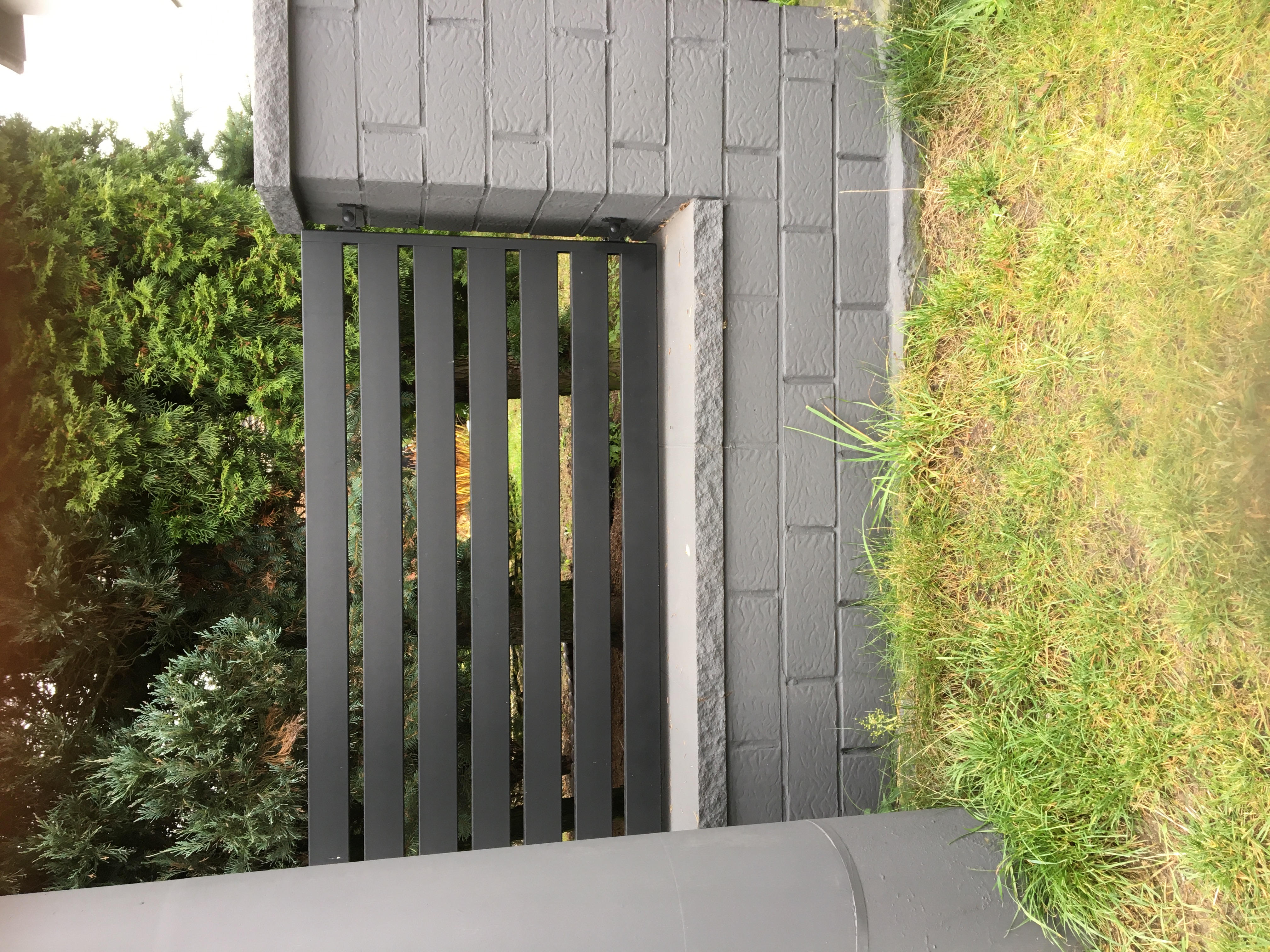 Nowoczesna, szara brama z pionowych, metalowych profili, osadzona w murowanym słupku z cegieł, widoczna z bliska na tle zieleni.