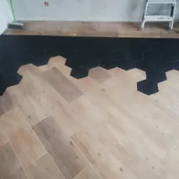 Układanie podłogi łączącej drewnopodobne panele i czarne heksagonalne płytki, widoczna przecinarka do płytek, drabina i miotła w tle.