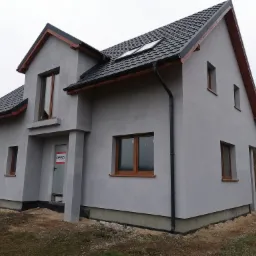 Ocieplony dom jednorodzinny z szarym styropianem, bez tynku, z brązowymi oknami i czarną dachówką, widok z zewnątrz na stan deweloperski.