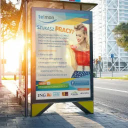 Przystanek autobusowy z plakatem reklamowym firmy Telmon, oferującej pracę konsultantom telefonicznym, w tle nowoczesny budynek biurowy i zieleń miejska.