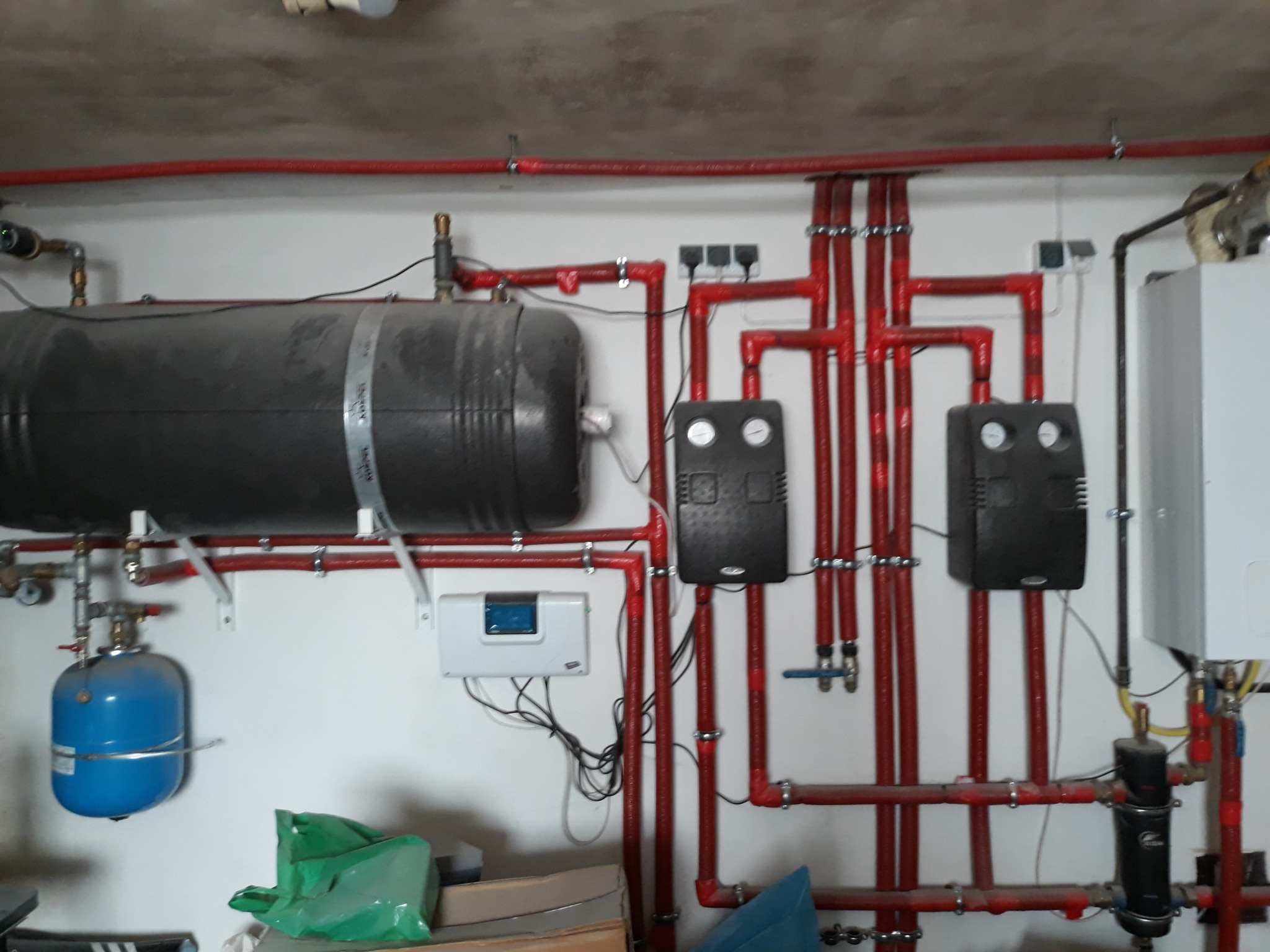 Kompleksowa instalacja grzewcza z czerwonymi rurami, zbiornikiem akumulacyjnym, rozdzielaczami i osprzętem na białej ścianie w piwnicy.