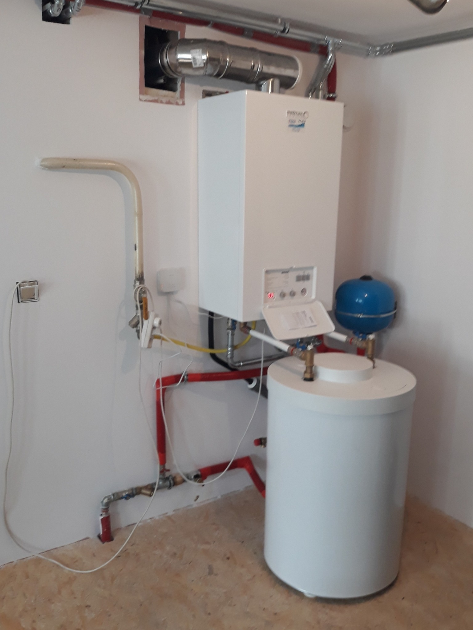 Biały piec gazowy kondensacyjny z niebieskim naczyniem wzbiorczym i czerwonymi rurami w nowym pomieszczeniu z surową podłogą, widoczne przewody wentylacyjne i instalacyjne.