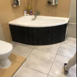 Narożna wanna obudowana czarnymi płytkami w łazience z beżowo-szarymi kaflami, widoczna toaleta i fragment osoby w skarpetkach.