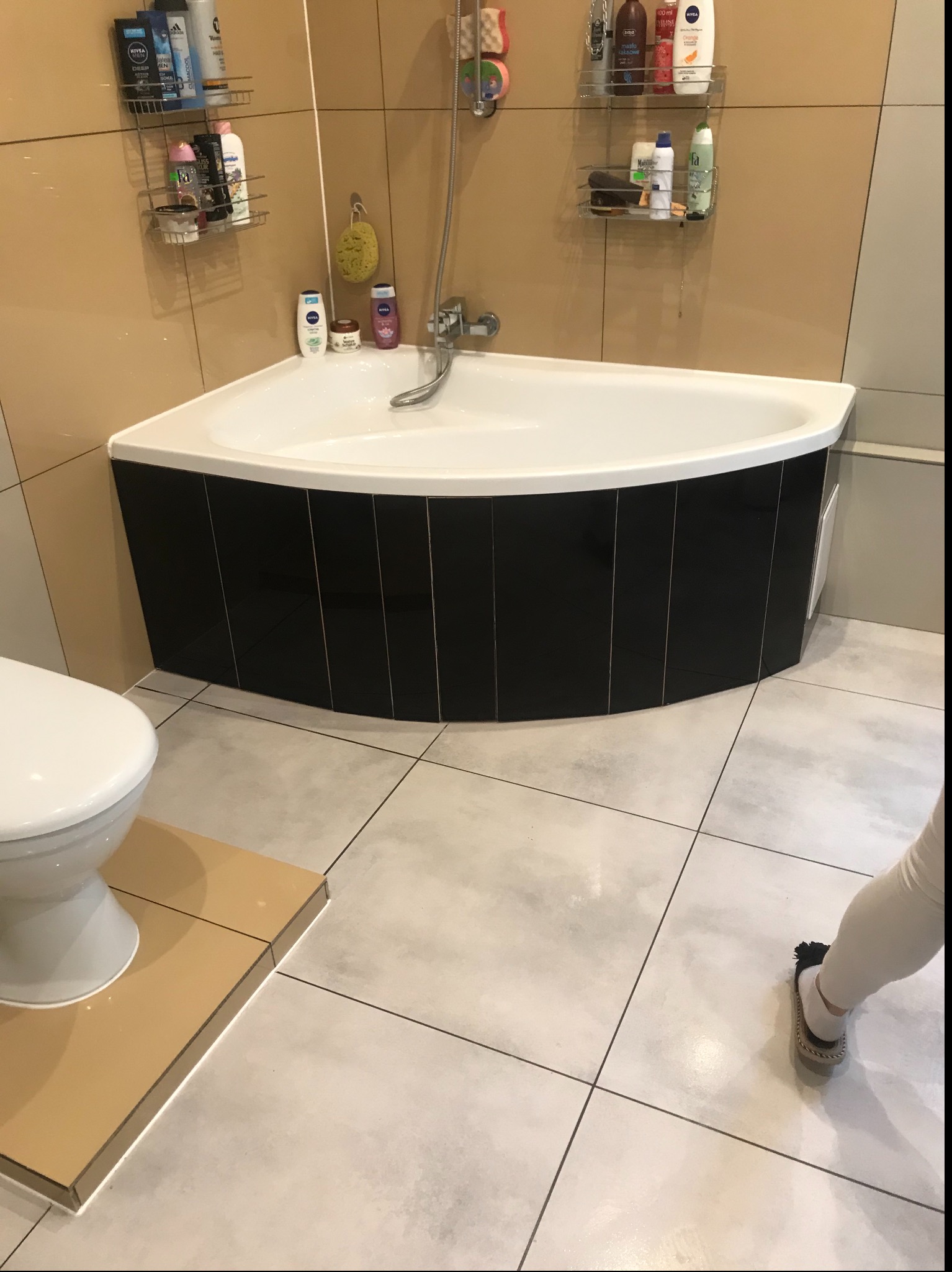 Narożna wanna obudowana czarnymi płytkami w łazience z beżowo-szarymi kaflami, widoczna toaleta i fragment osoby w skarpetkach.