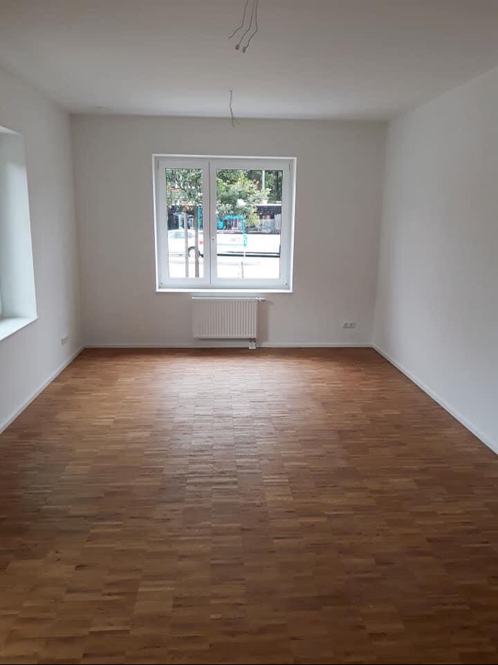 Pusty pokój po remoncie z parkietem w jodełkę, białymi ścianami i oknem z widokiem na zieleń, widoczne kable wystające z sufitu.