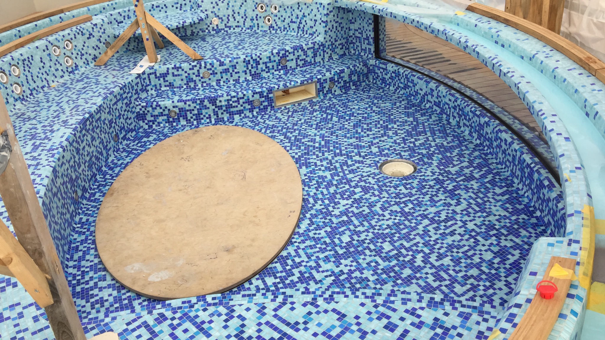 Wnętrze mozaikowej wanny jacuzzi w trakcie budowy, widoczne drewniane podpory, okrągła pokrywa i otwory na dysze.