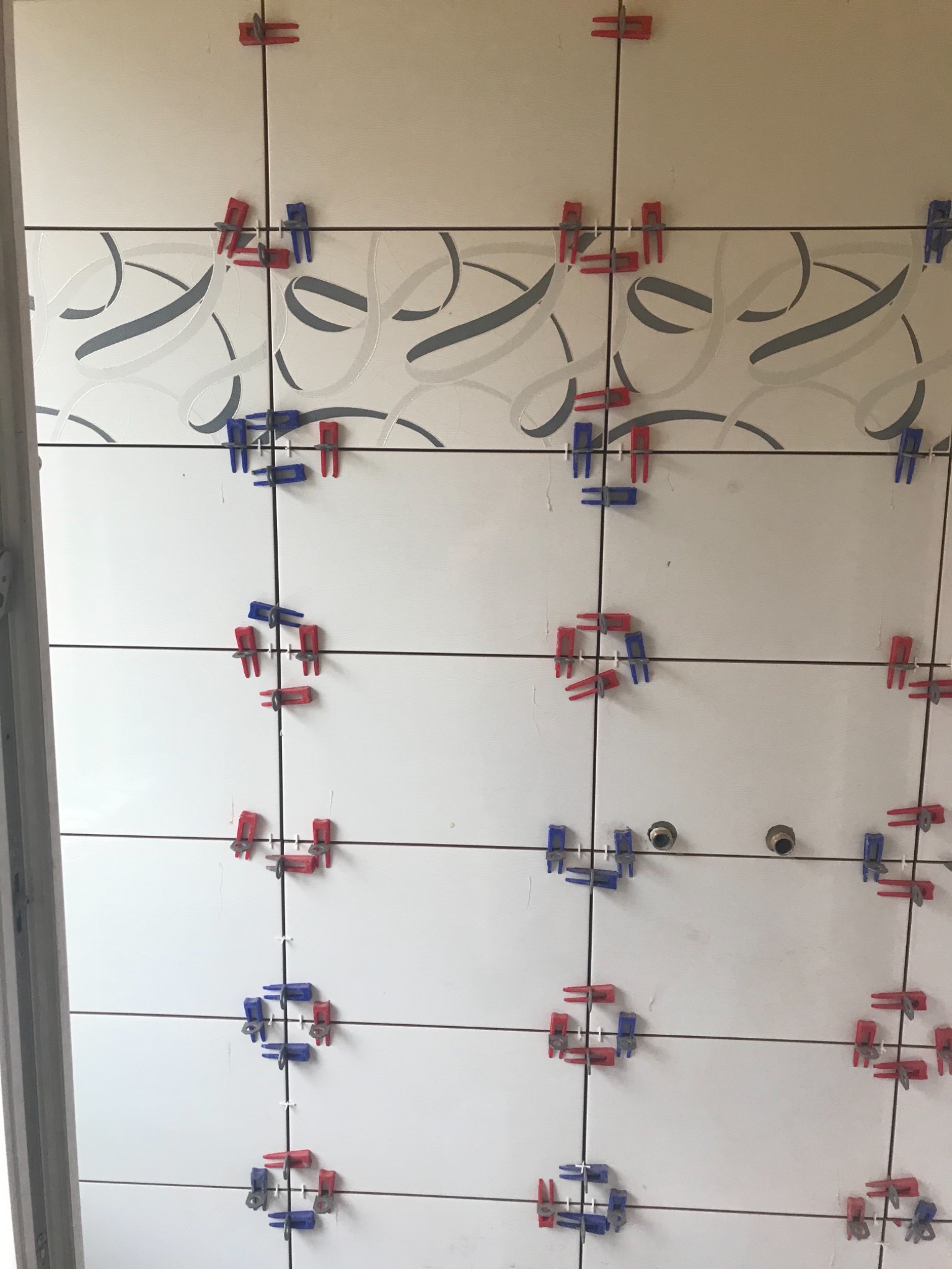 Białe płytki z dekoracyjnym wzorem w trakcie układania, z użyciem czerwono-niebieskich klipsów poziomujących, widoczne otwory na instalacje.