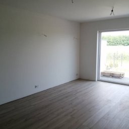 SEBUD Sebastian Przyłucki - Surowe wnętrze pokoju z nową podłogą z paneli, gładkimi, białymi ścianami z wystającymi przewodami i widokiem na zewnątrz przez duże okno.