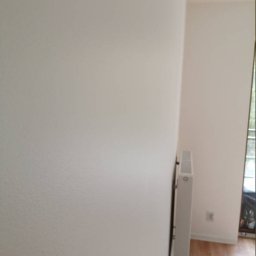 SEBUD Sebastian Przyłucki - Świeżo pomalowana, jasna ściana w rogu pokoju z widocznym grzejnikiem i fragmentem podłogi z paneli.