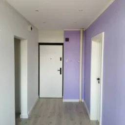 Korytarz w Puszczykowie po remoncie, z pomalowanymi na fioletowo ścianami, drewnopodobną podłogą i białymi drzwiami; widoczne przygotowania do montażu oświetlenia sufitowego.