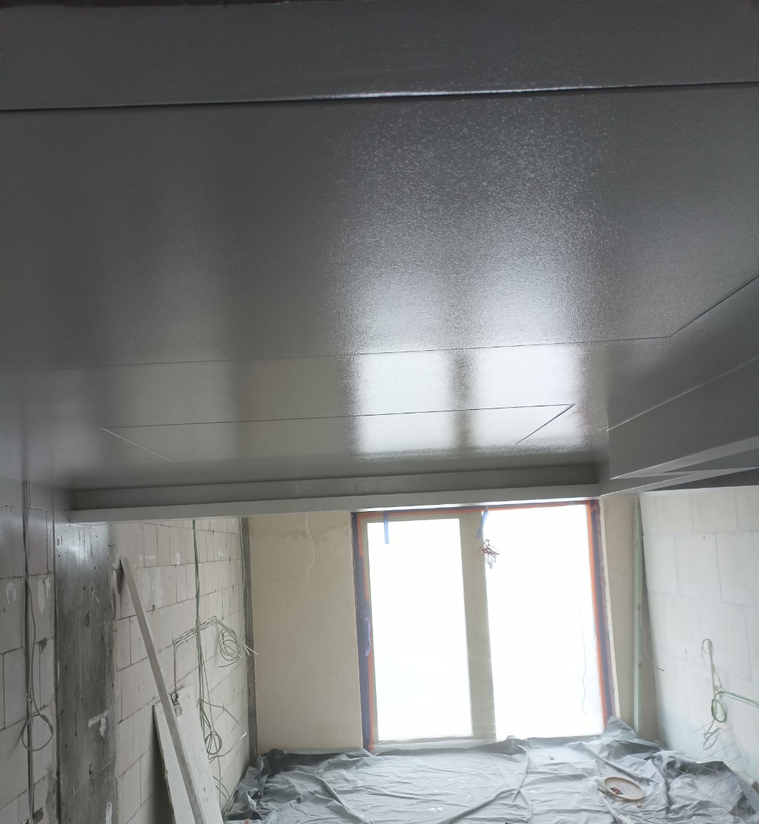 Wnętrze w trakcie prac wykończeniowych: świeżo pomalowany sufit na szaro, okno zabezpieczone folią, ściany z widocznymi przewodami elektrycznymi.