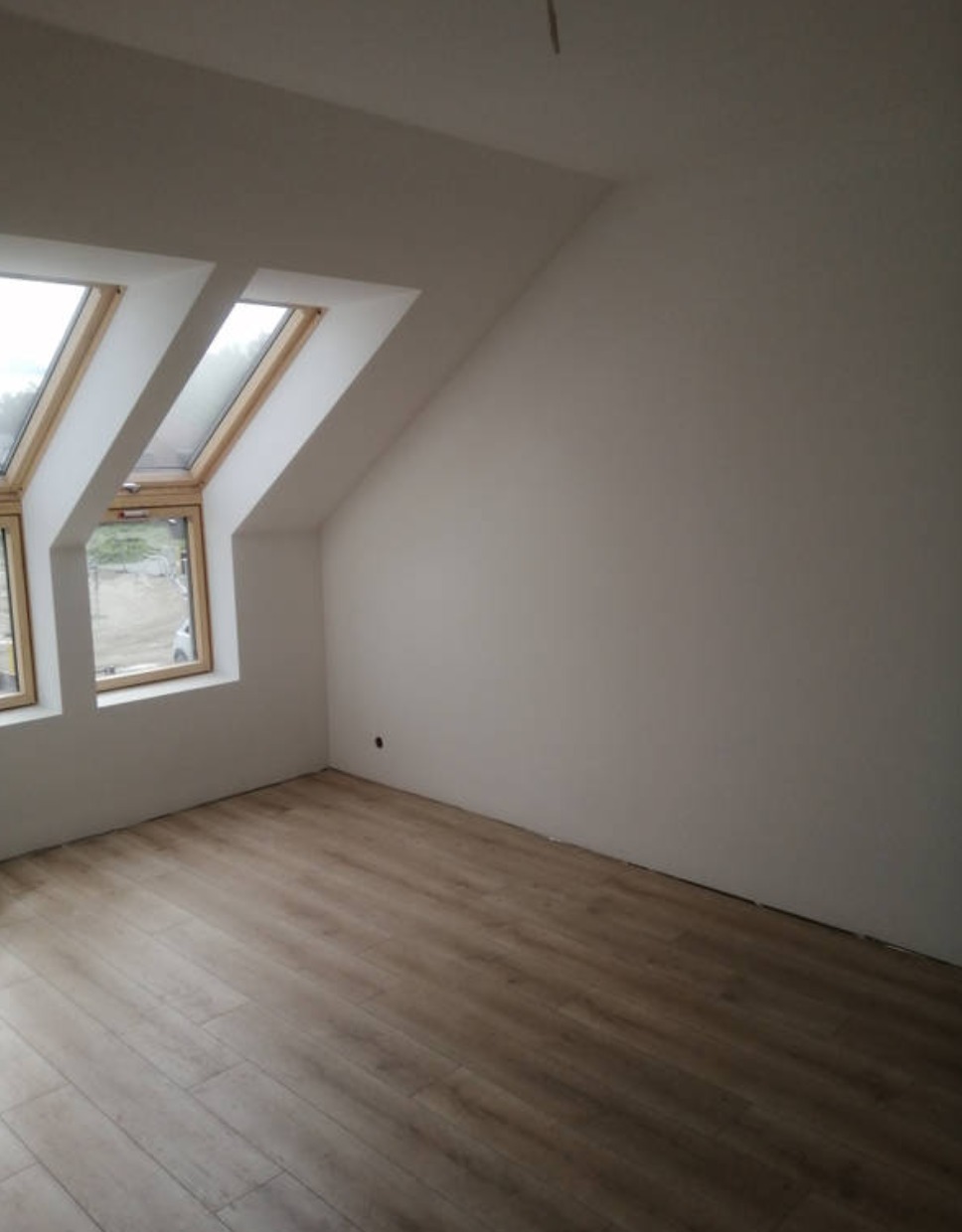 Poddasze z dwoma oknami dachowymi, wykończone panelami podłogowymi i świeżo pomalowanymi ścianami na biało, widoczny fragment wystającego kabla z sufitu.