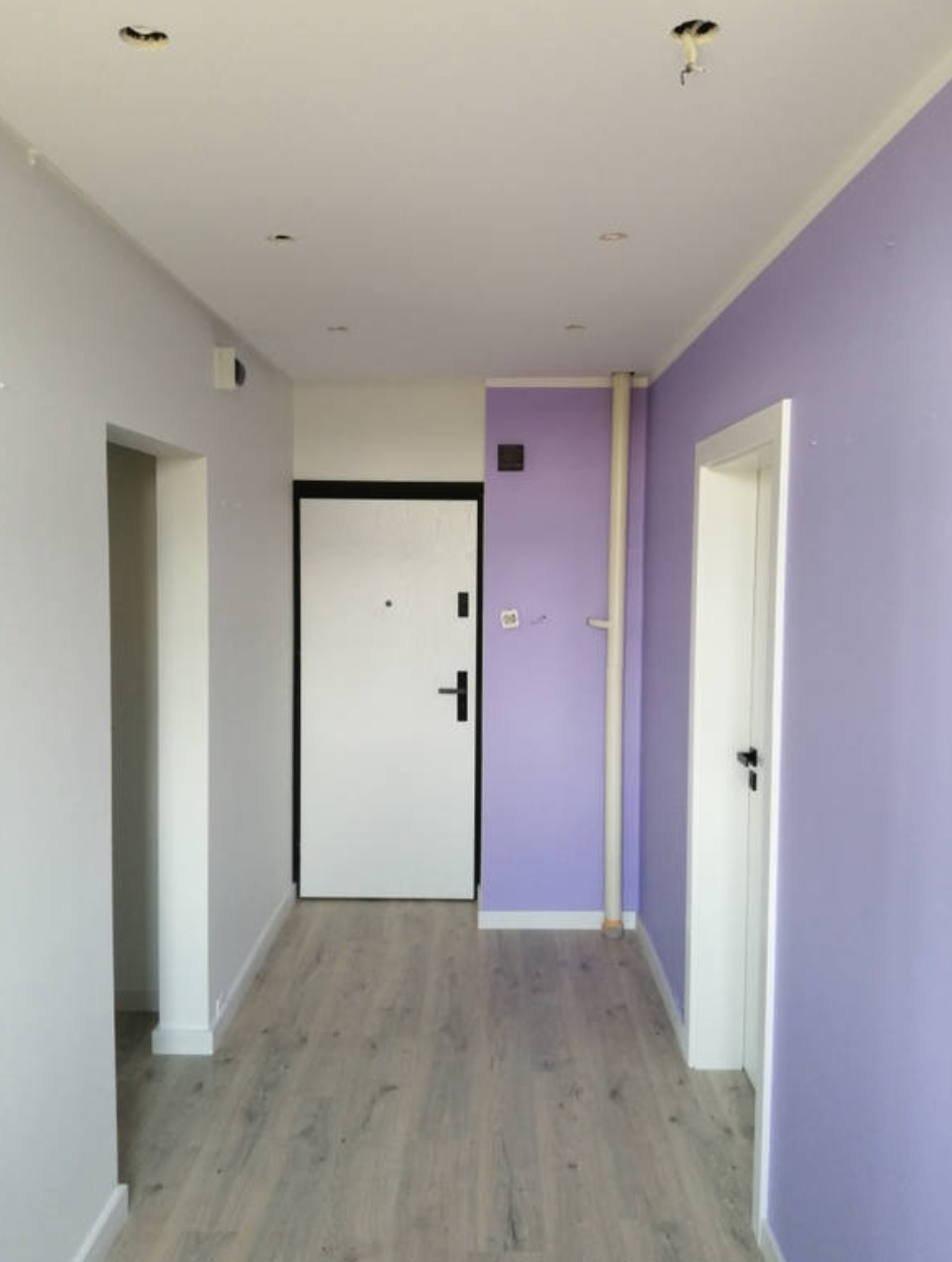 Korytarz w Puszczykowie po remoncie, z pomalowanymi na fioletowo ścianami, drewnopodobną podłogą i białymi drzwiami; widoczne przygotowania do montażu oświetlenia sufitowego.