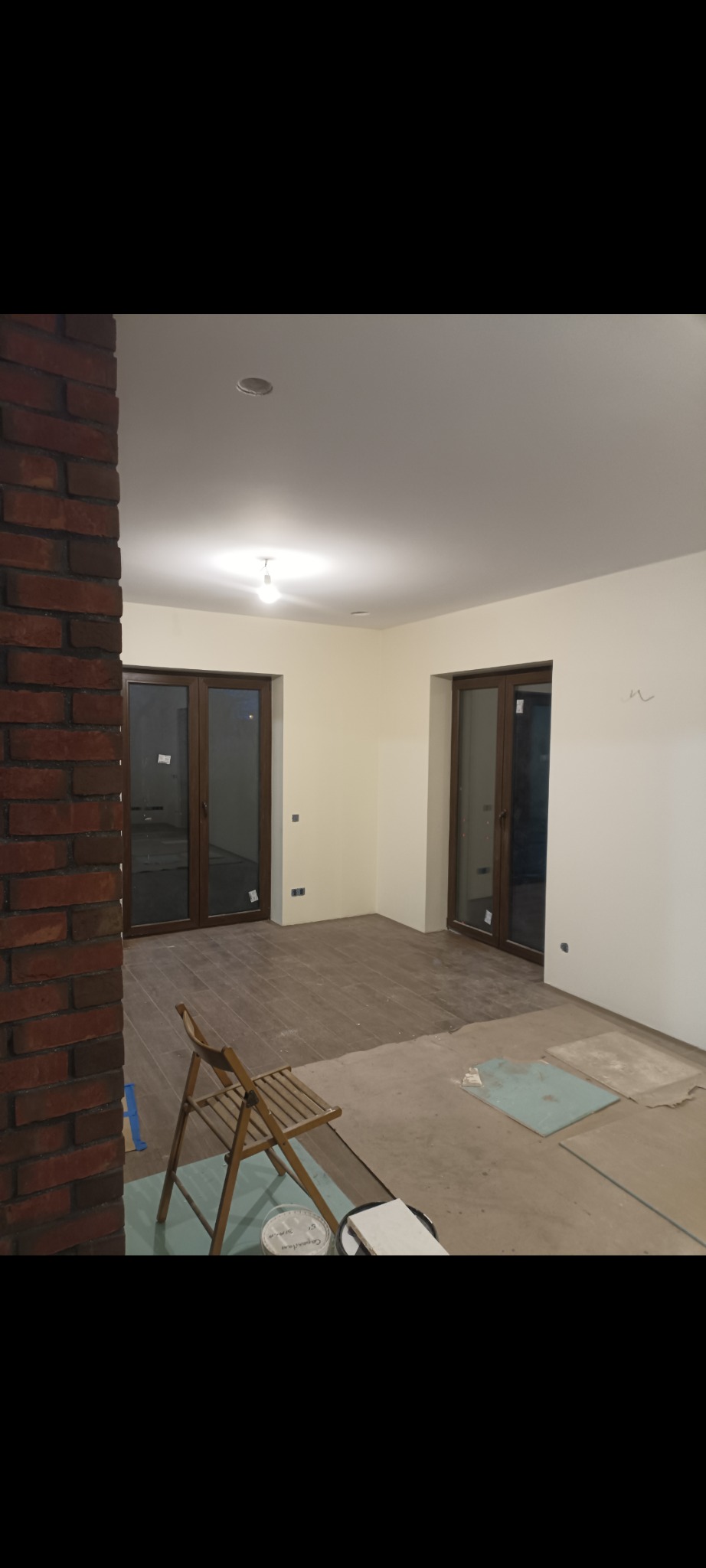 Wnętrze w trakcie prac: świeżo pomalowane ściany w odcieniach bieli, zabezpieczona podłoga, widoczny fragment ściany z cegły, drabina malarska i puszka farby.